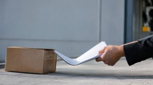 Une main en costume retire un document d'un carton posé au sol.